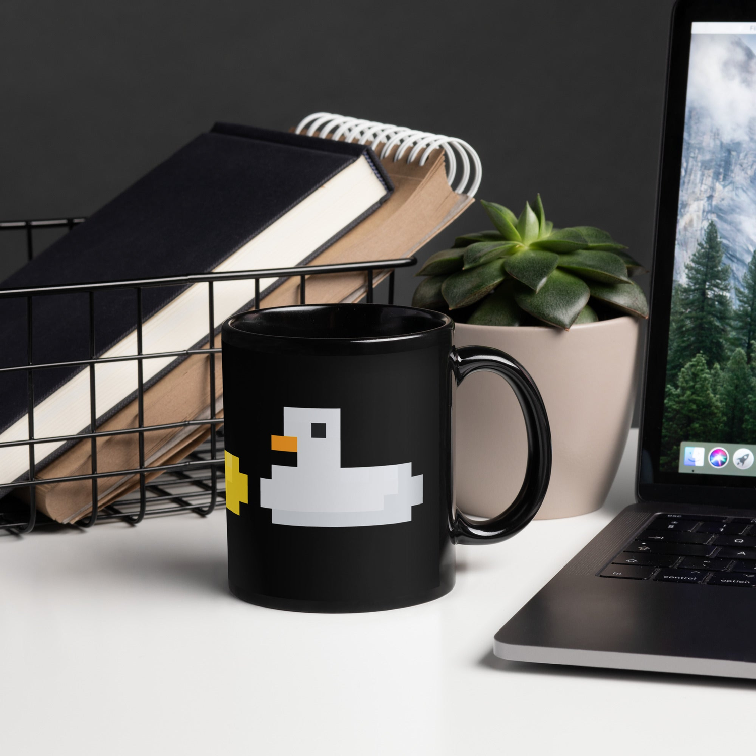 A black mug featuring a pixel art gray duck and partial yellow duck sits on a modern white desk. Surrounding it are a wire basket of books, a spiral notebook, a potted succulent, and an open laptop.
