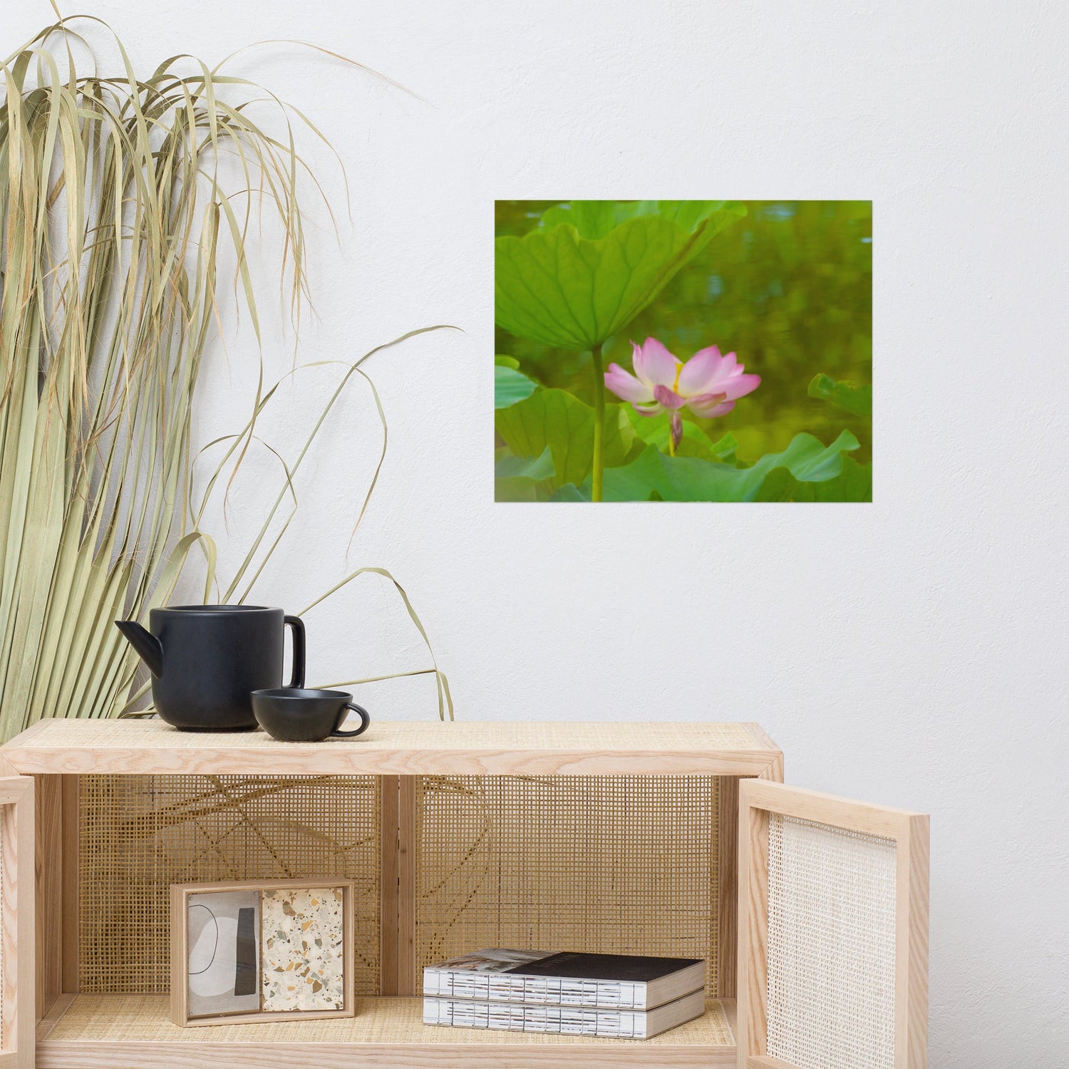 A soft-focus lotus flower print hangs on a white wall above a natural wood and cane cabinet. The serene scene features a pink lotus bloom and lush green leaves. A black teapot and cup sit on top, with dried grass decor adding a calming, organic touch.