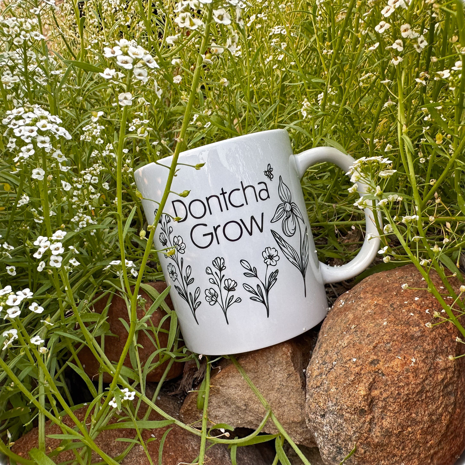 A white ceramic mug with black line art of wildflowers and the phrase “Dontcha Grow” rests on rocks surrounded by blooming white flowers and lush green grass, blending naturally into the garden setting.