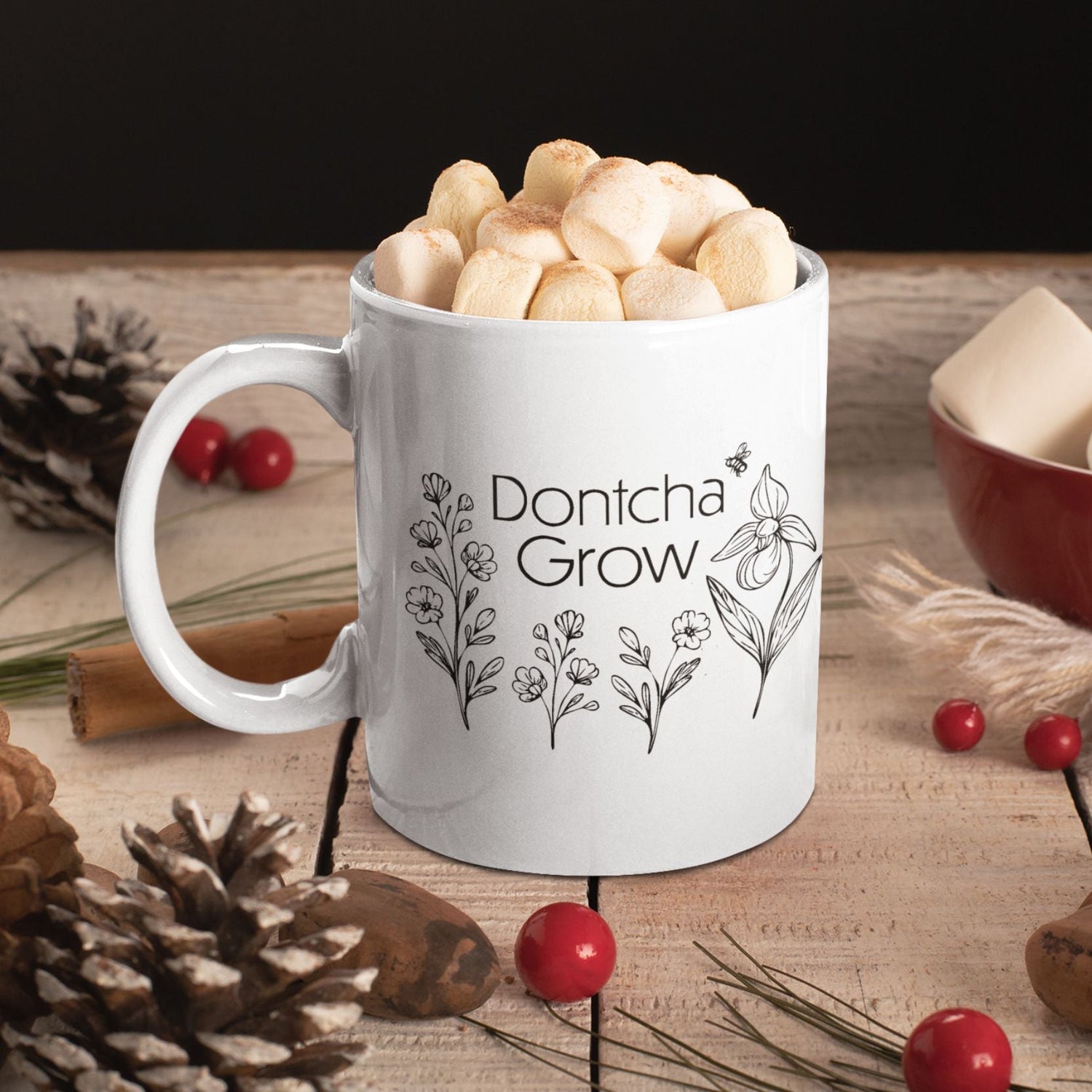 A white “Dontcha Grow” floral mug is filled with hot cocoa and toasted marshmallows, surrounded by festive holiday decor on a rustic wood table, creating a cozy winter scene.