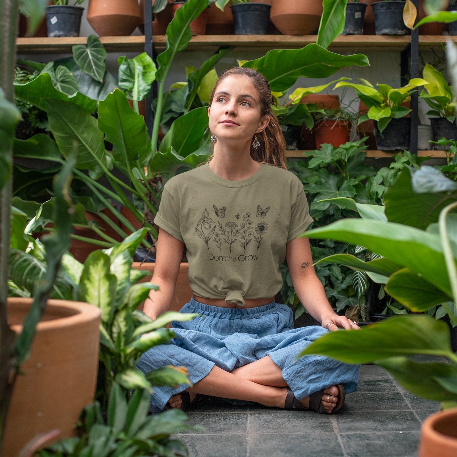 A woman sits cross-legged in a lush greenhouse wearing an olive green knotted t-shirt with black line art of wildflowers, butterflies, and the phrase “Dontcha Grow.” She's surrounded by potted plants, appearing peaceful and thoughtful.