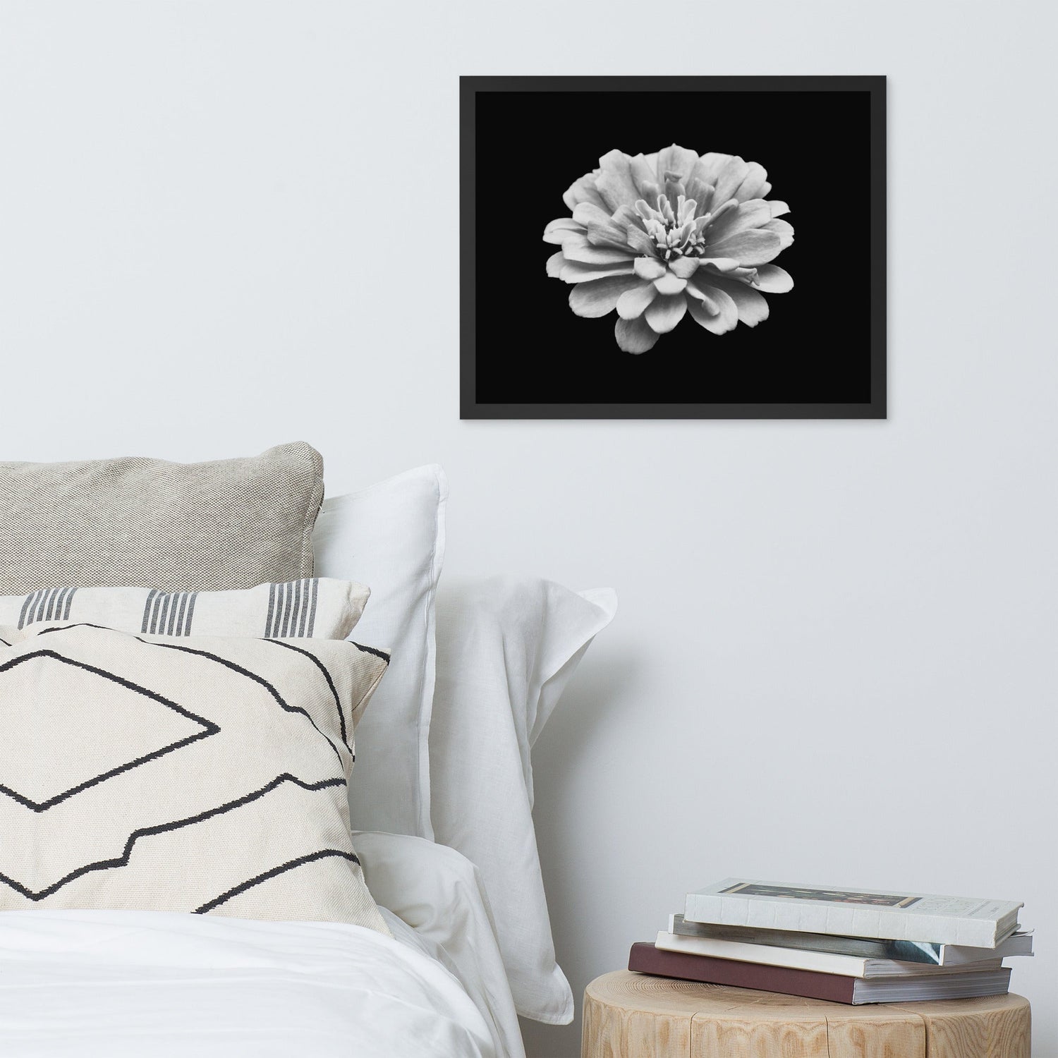 A black and white framed photo of a zinnia flower hangs above a neatly made bed with neutral pillows and white bedding. A round wooden nightstand holds a small stack of books, completing the clean, modern bedroom aesthetic.